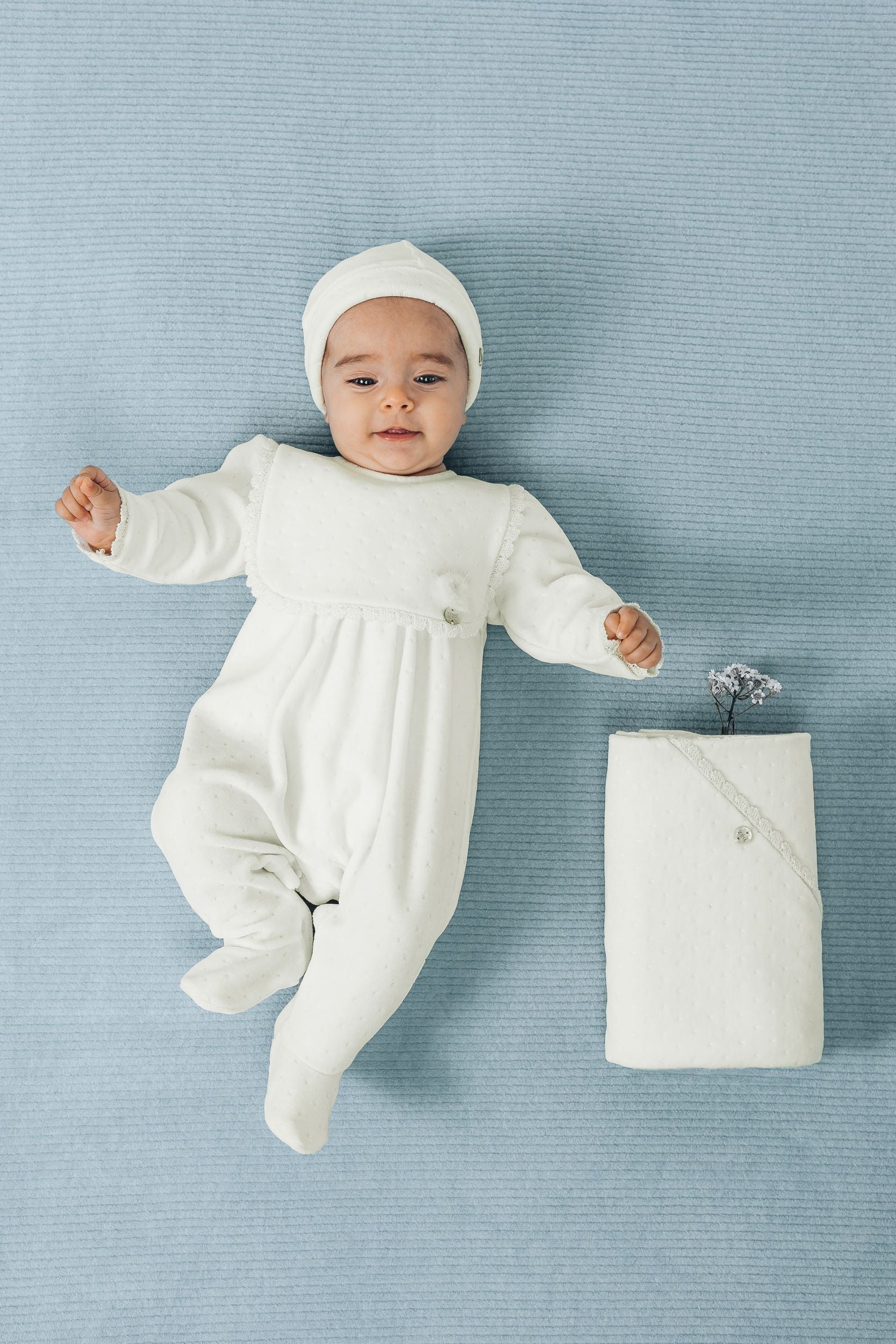A baby is wearing a white onesie with a lace collar;A baby in a white outfit is laying on a blue blanket.