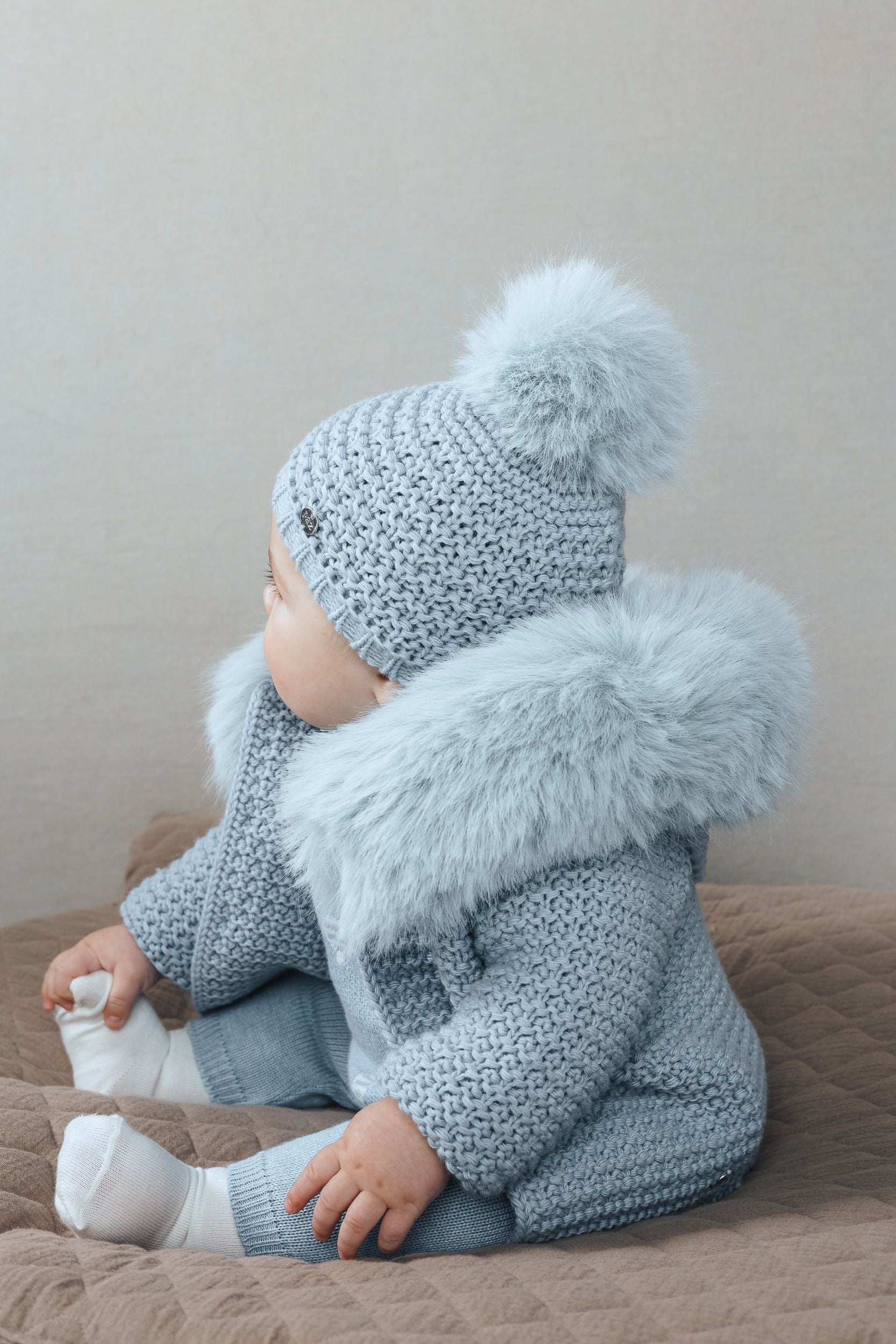 A baby is wearing a blue sweater with a fur collar.;A baby is wearing a blue sweater with a fur hood.;A baby is wearing a pink sweater with a fur collar;A baby is sitting on a bed wearing a hat and a jacket.