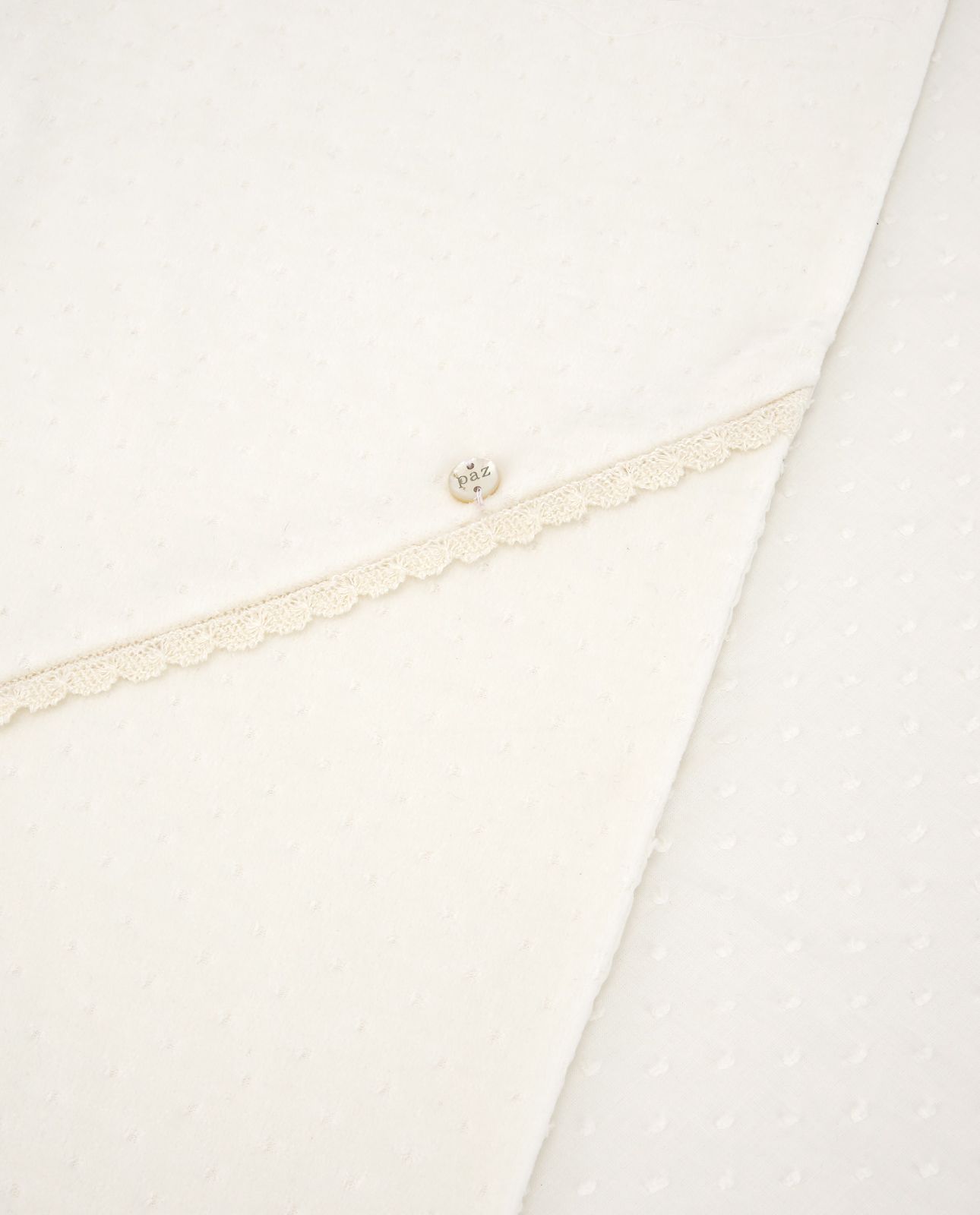 A white blanket with a pearl embroidered border is laying on a white surface.;A close up of a white cloth with a lace trim and a button.