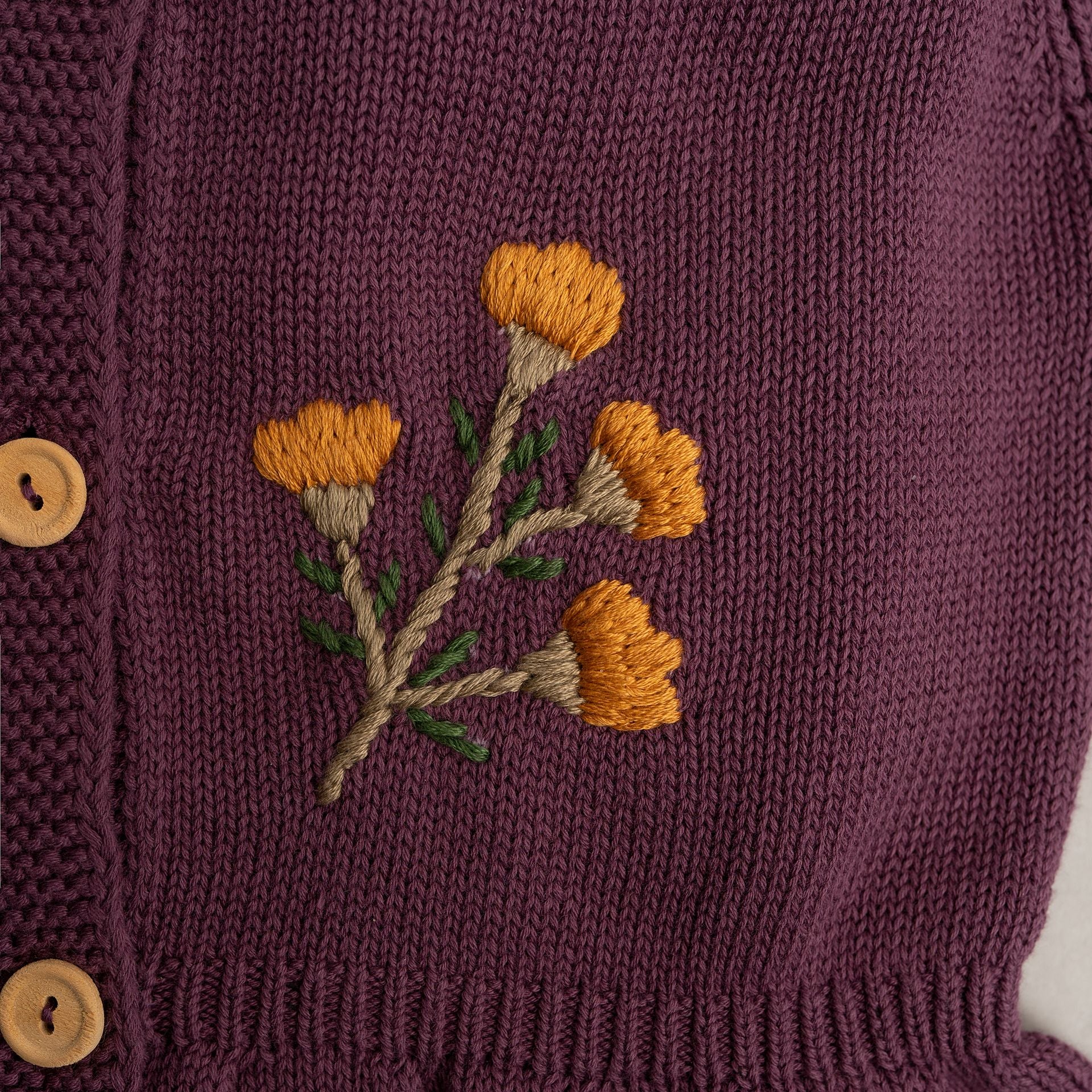 A baby girl is wearing a purple cardigan with flowers embroidered on it.;A purple sweater with ruffles on the bottom is on a white background.;A close up of a purple sweater with a flower embroidered on it.;A close up of a purple sweater with yellow flowers embroidered on it.