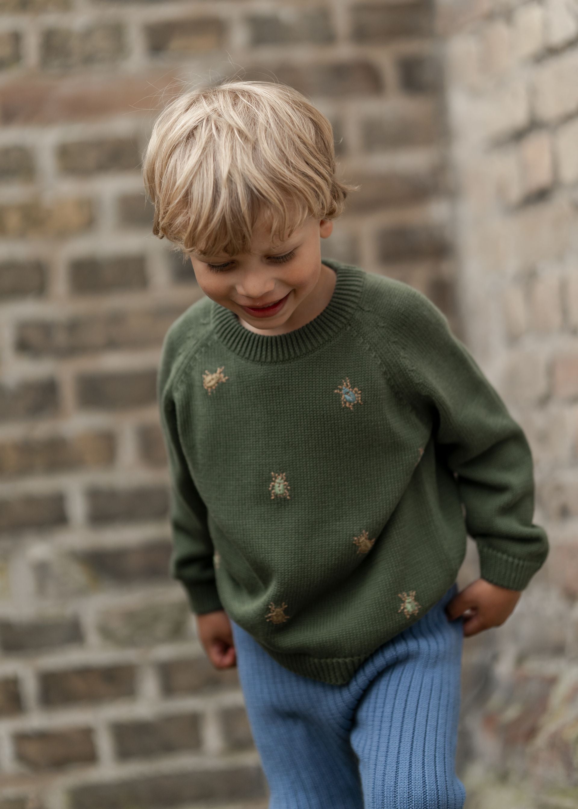 A young boy wearing a green sweater and blue pants is standing in front of a brick wall.;A green sweater with gold bees embroidered on it;A green sweater with long sleeves on a white background;A close up of a green sweater with bees embroidered on it.
