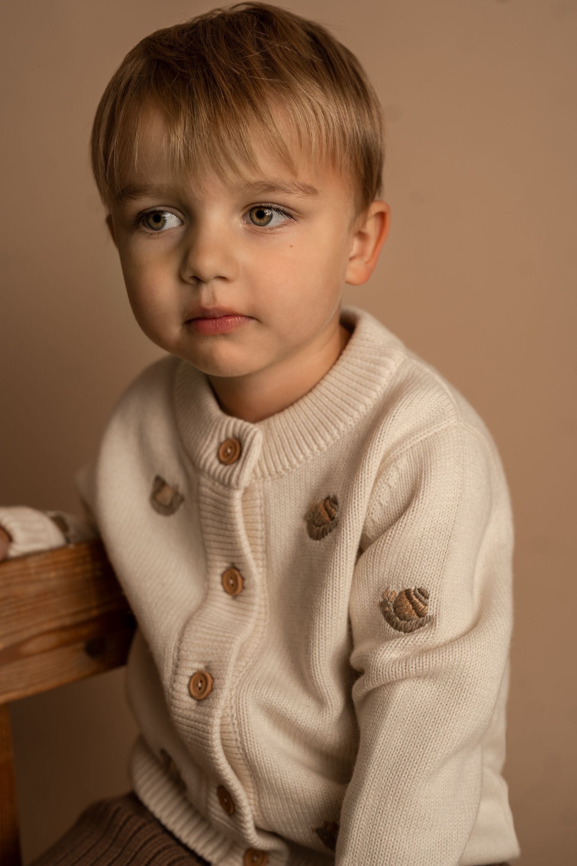 A young boy is sitting on a chair wearing a white sweater.;A white cardigan with squirrels embroidered on it;The back of a white sweater with long sleeves on a white background.;A close up of a sweater with a sheep embroidered on it.