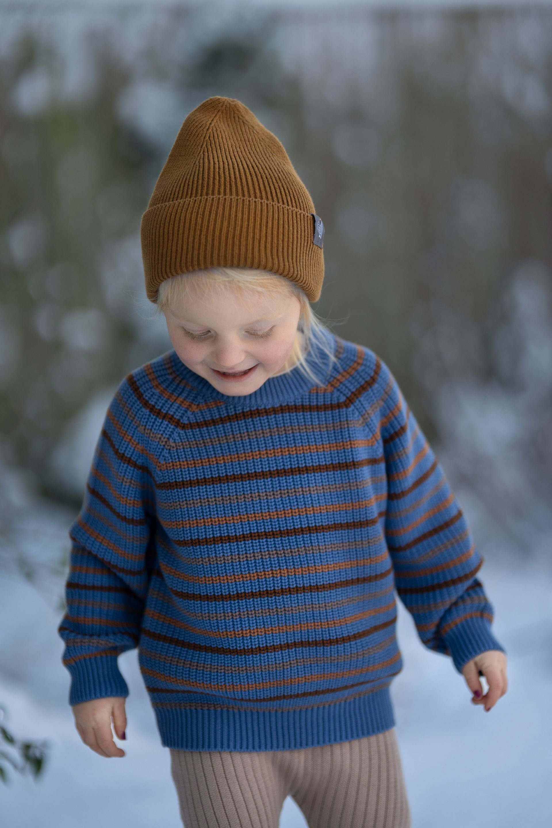 A little girl wearing a striped sweater and a brown hat is standing in the snow.;A blue and brown striped sweater with long sleeves on a white background.;A blue and orange striped sweater with long sleeves on a white background.