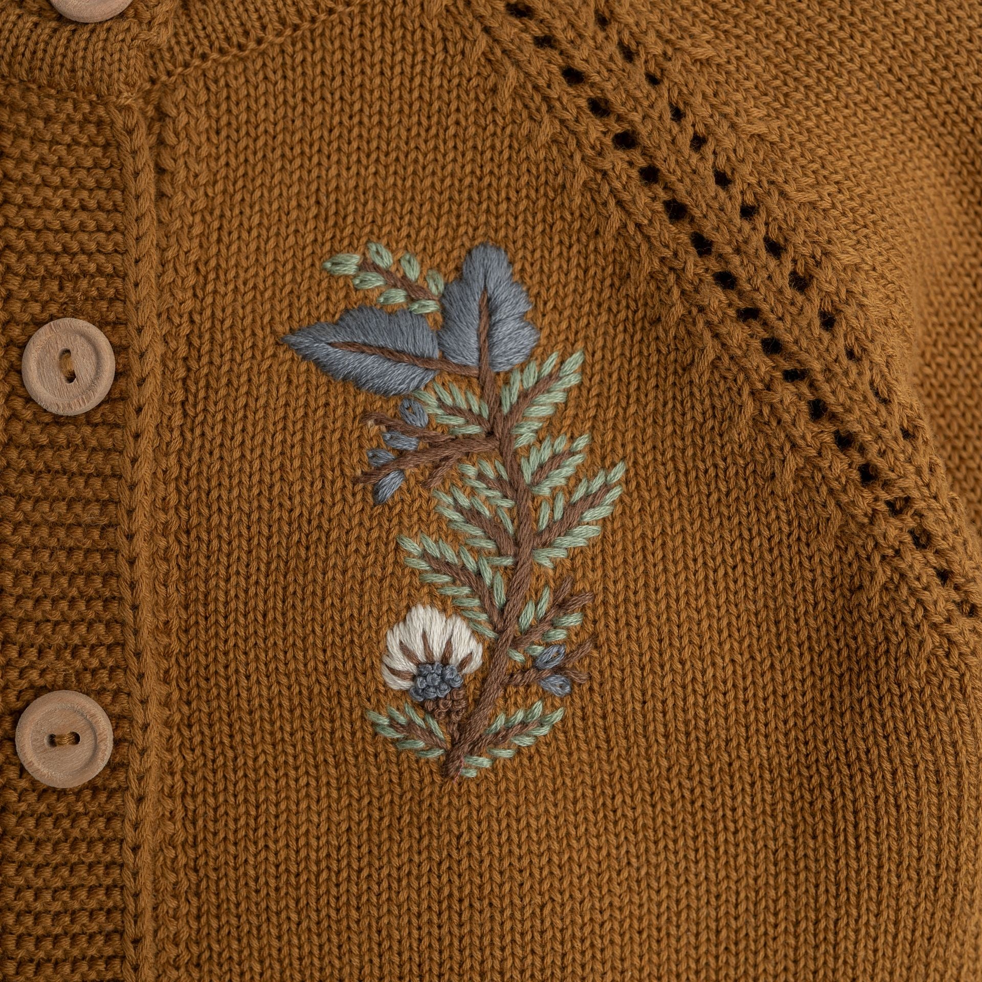A little girl wearing a brown sweater and a white hat is standing in the snow.;A baby girl is wearing a brown cardigan with embroidery on it.;A close up of a sweater with a floral embroidery on it.;A close up of a sweater with a flower embroidered on it.
