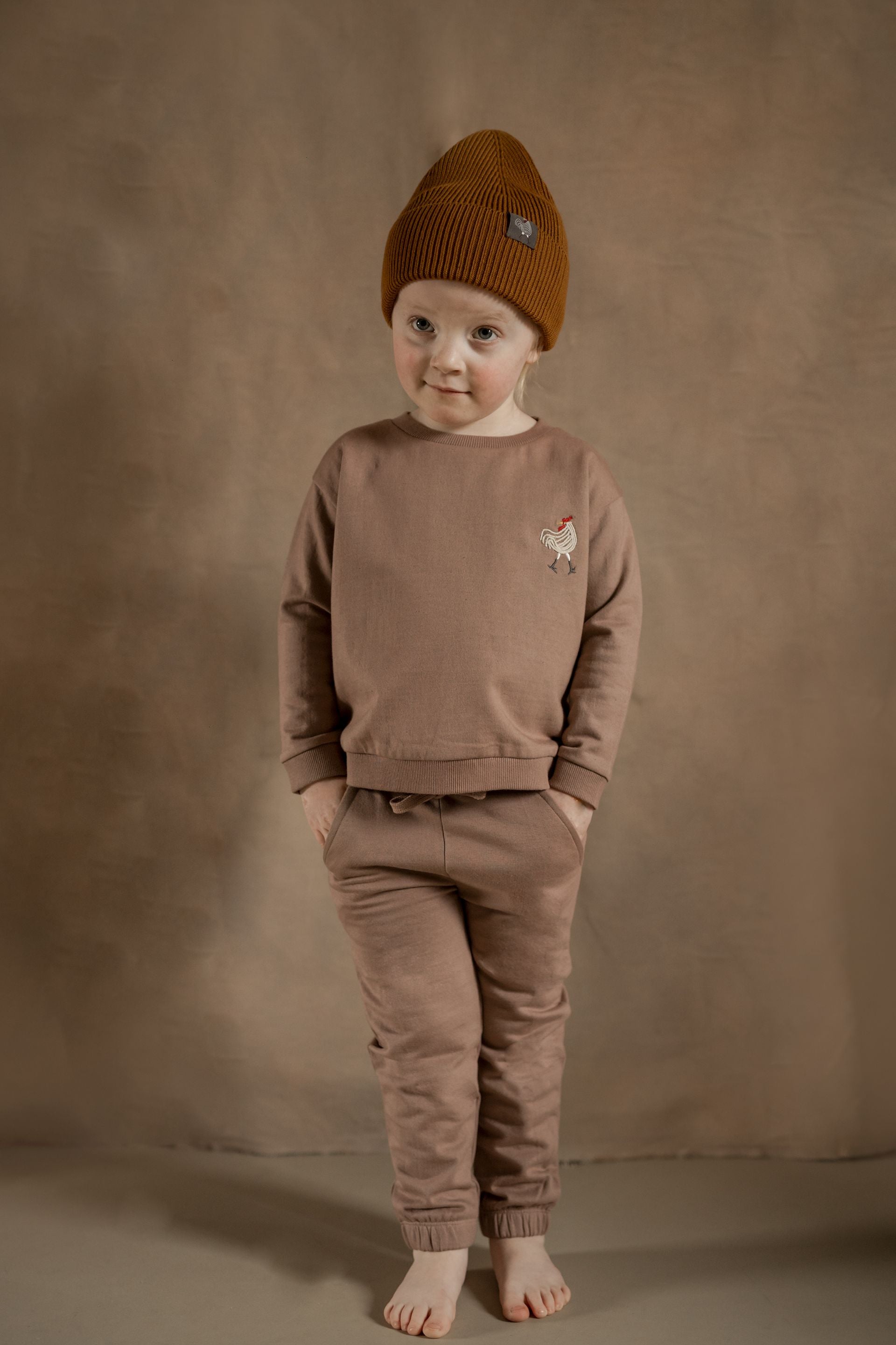 A little boy wearing a brown hat and a brown sweater is standing with his hands in his pockets.;A pair of brown sweatpants with a sheep embroidered on the back pocket;A pair of brown sweatpants on a white background