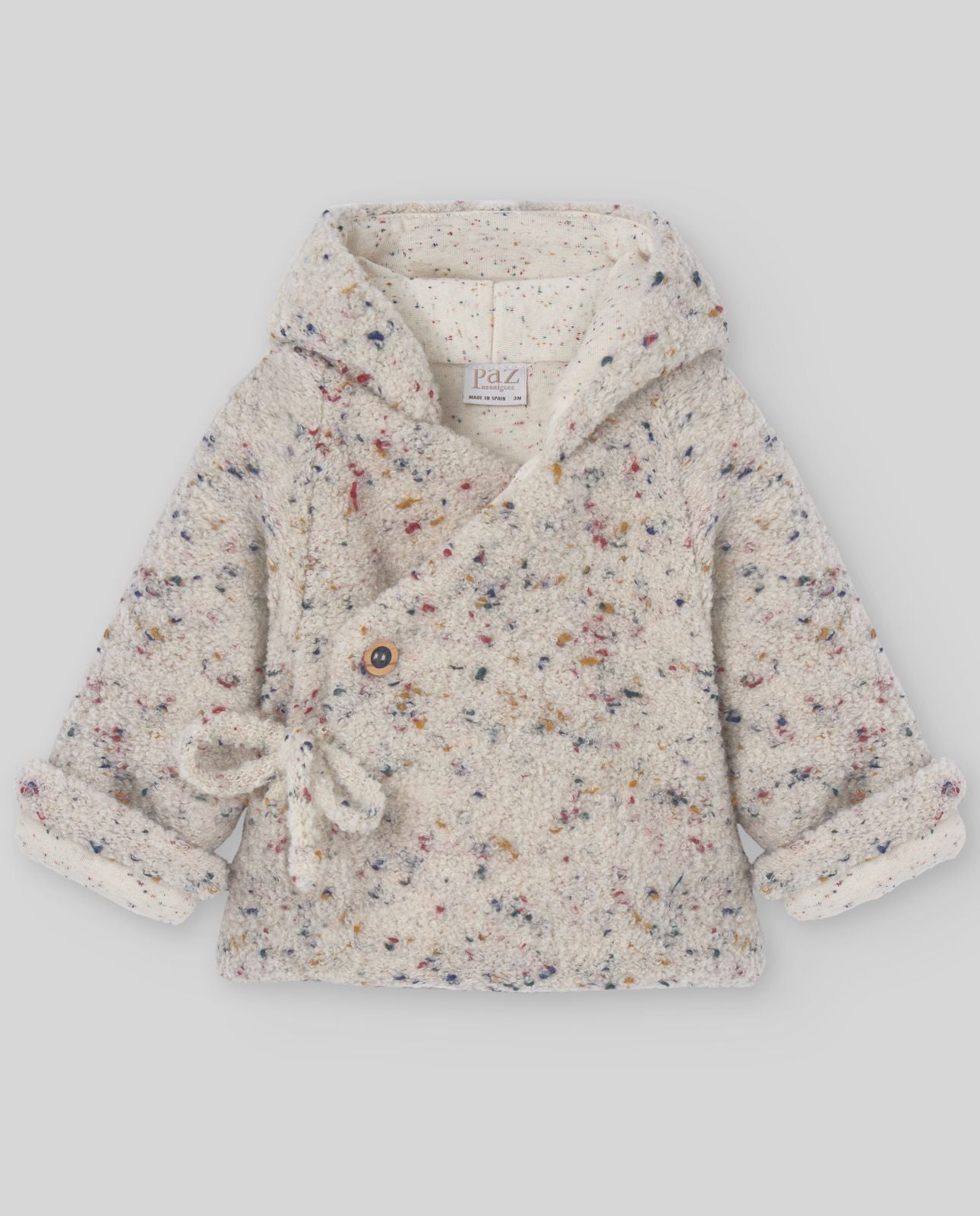 A baby is wearing a white jacket with a hood.;The back of a white jacket with a blue pom pom on the hood.